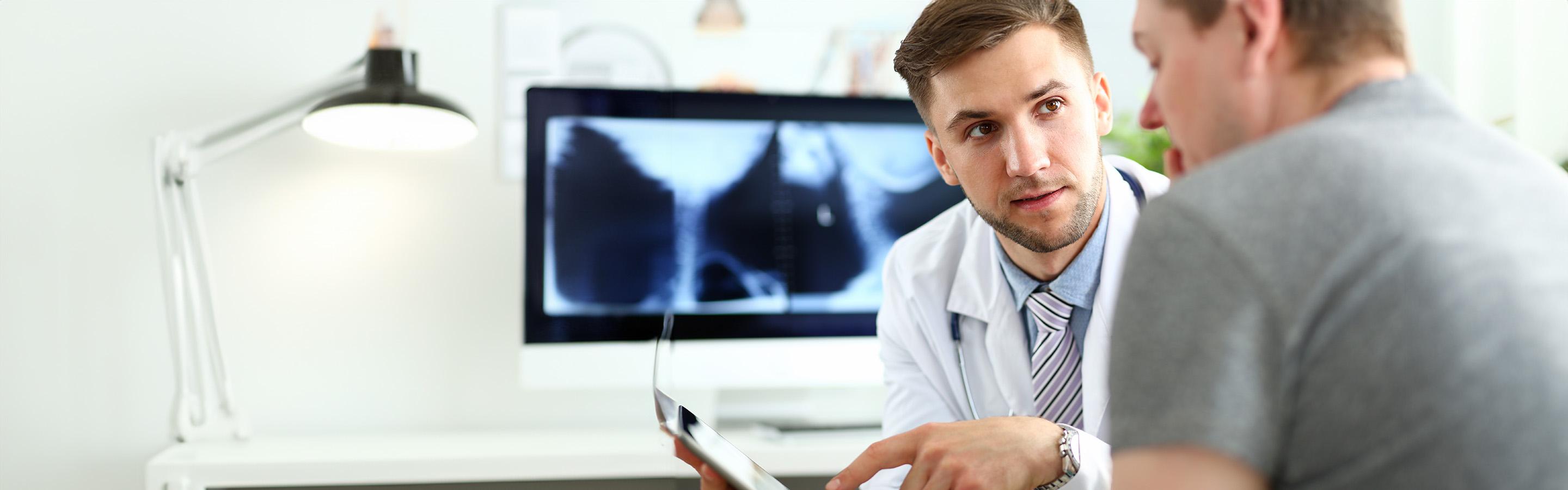 A male caregiver and a patient have a discussion in a clinical setting, x-ray of spine visible in background