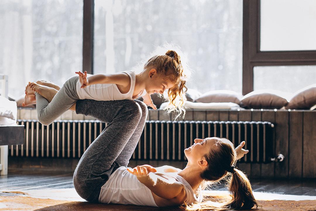 Mother lying on her back in a yoga pose with her daughter resting on her legs