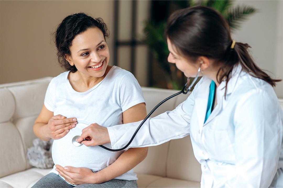 Woman sitting on a couch, holding her stomach while a doctor examines her with a stethoscope